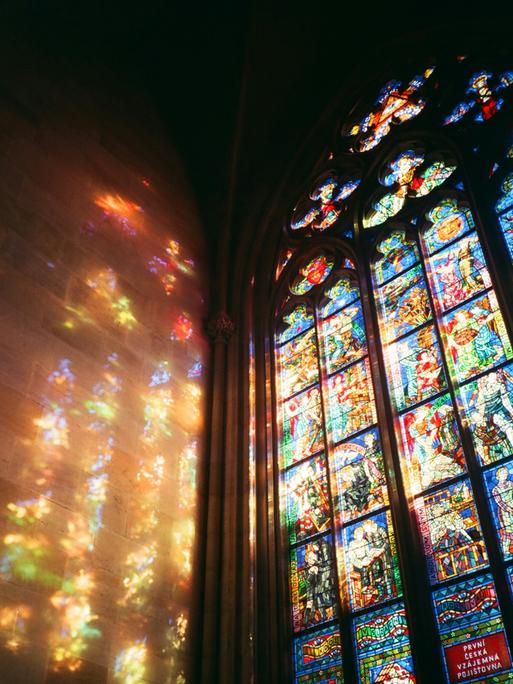 Licht scheint durch ein buntes Prager Kirchenfester und reflektiert farbig an der Wand