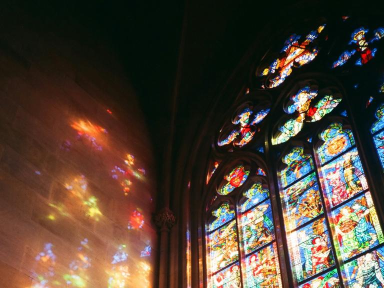 Licht scheint durch ein buntes Prager Kirchenfester und reflektiert farbig an der Wand
