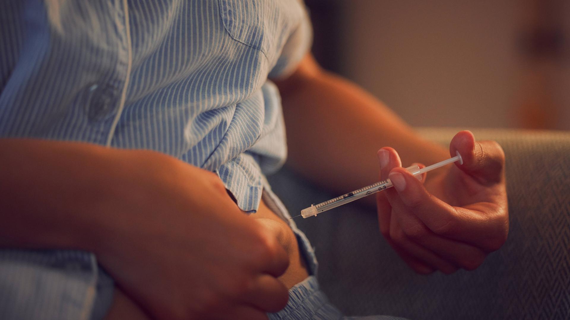 Eine Person setzt eine Spritze am Bauch an. Es ist ein Insulin-Spritze. Die Person trägt einen Pyjama.