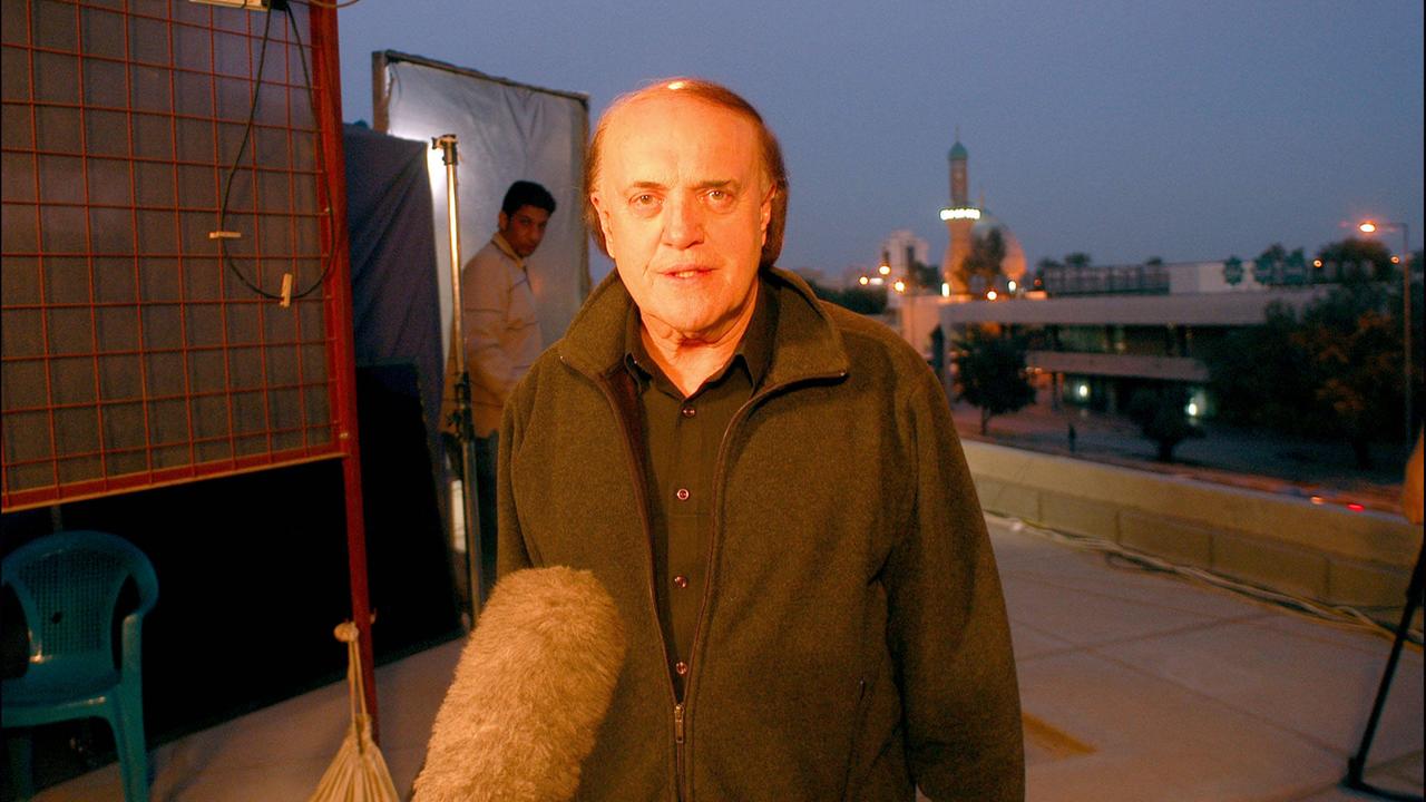 Peter Arnett steht auf einem Hausdach und spricht in ein Mikrofon. 