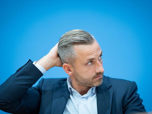 Ulrich Siegmund, im Sakko vor blauem Hintergrund, schaut zur Seite und greift sich an den Hinterkopf. Ulrich Siegmund, im Sakko vor blauem Hintergrund, schaut zur Seite und greift sich an den Hinterkopf.