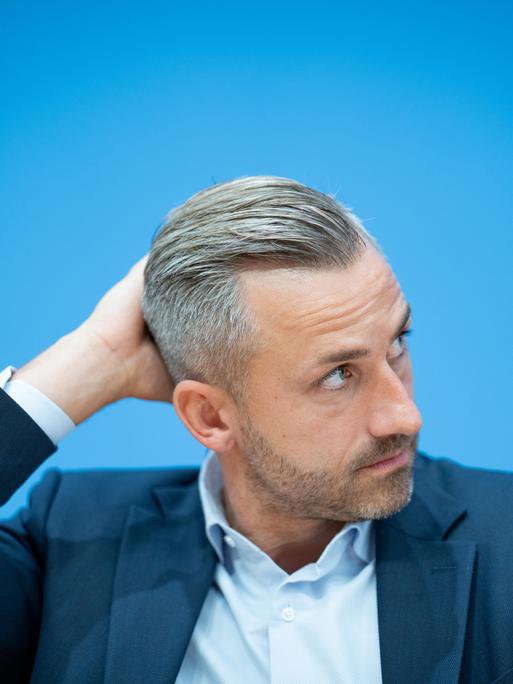 Ulrich Siegmund, im Sakko vor blauem Hintergrund, schaut zur Seite und greift sich an den Hinterkopf. Ulrich Siegmund, im Sakko vor blauem Hintergrund, schaut zur Seite und greift sich an den Hinterkopf.