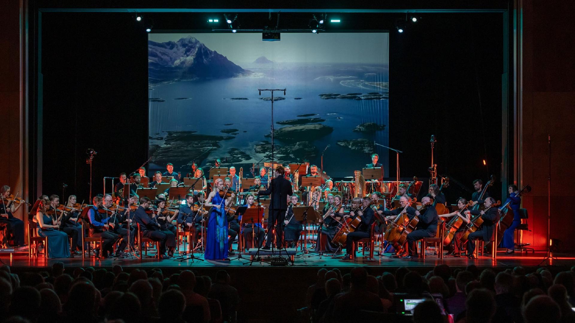Blick auf das Orchester, das mit Geigerin und Dirigent vor einer Videowand spielt, auf dem arktische Weiten zu sehen sind. Blick auf das Orchester, das mit Geigerin und Dirigent vor einer Videowand spielt, auf dem arktische Weiten zu sehen sind.