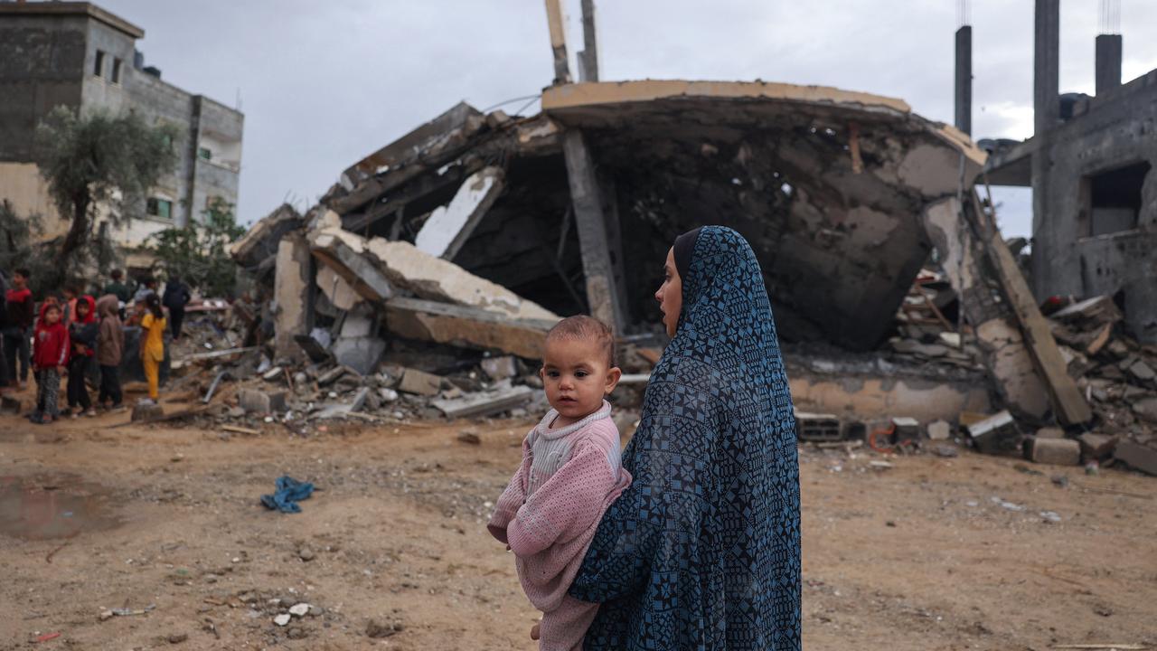 Eine Frau steht mit einem Kleinkind auf dem Arm vor den Trümmern zerstörter Gebäude. Im Hintergrund eine Gruppe Kinder.