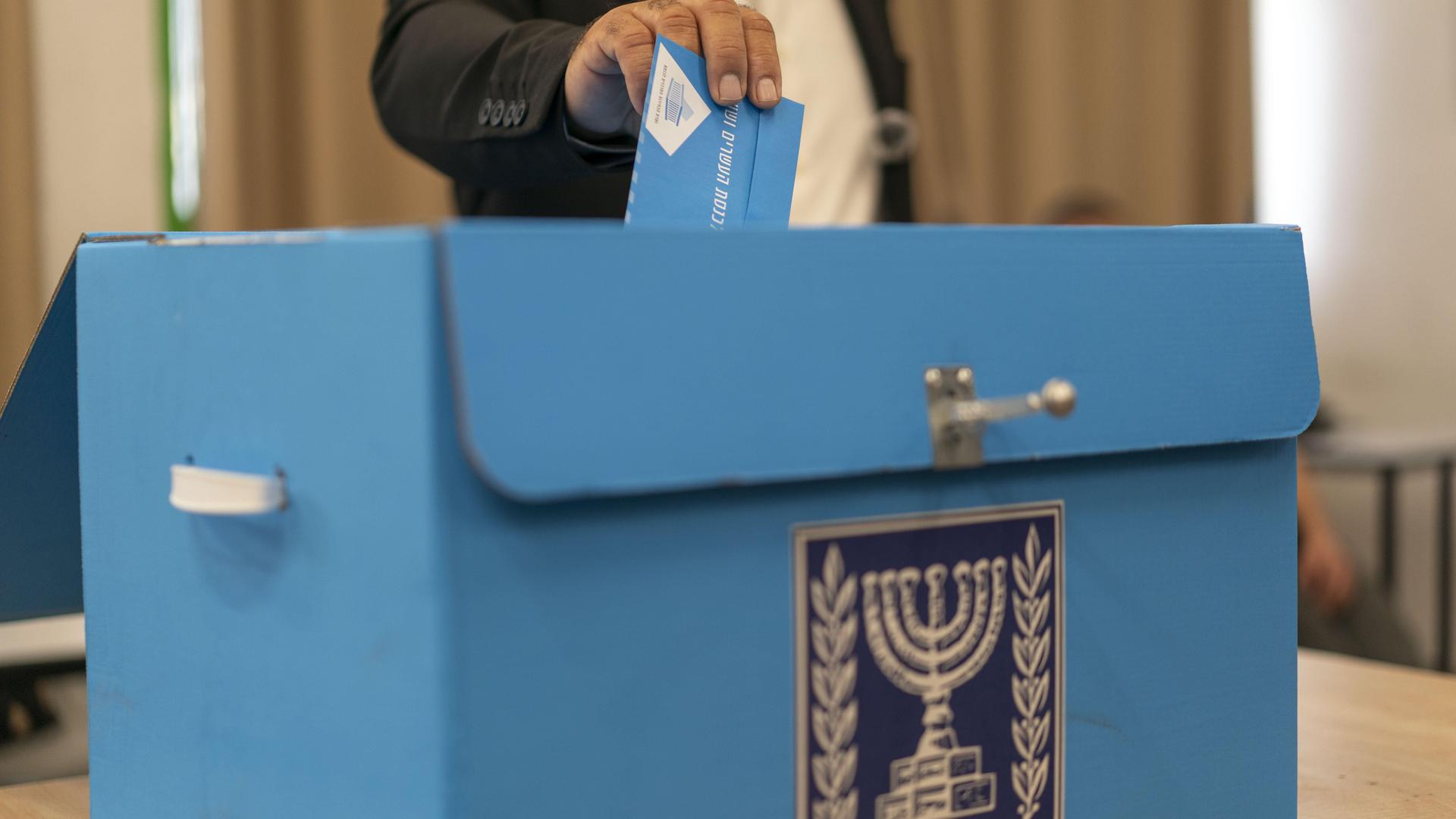 Ein Mann steckt seinen Wahlumschlag in eine Wahlkiste, auf der der siebenarmige Leuchter zu sehen ist. Ein Mann steckt seinen Wahlumschlag in eine Wahlkiste, auf der der siebenarmige Leuchter zu sehen ist.