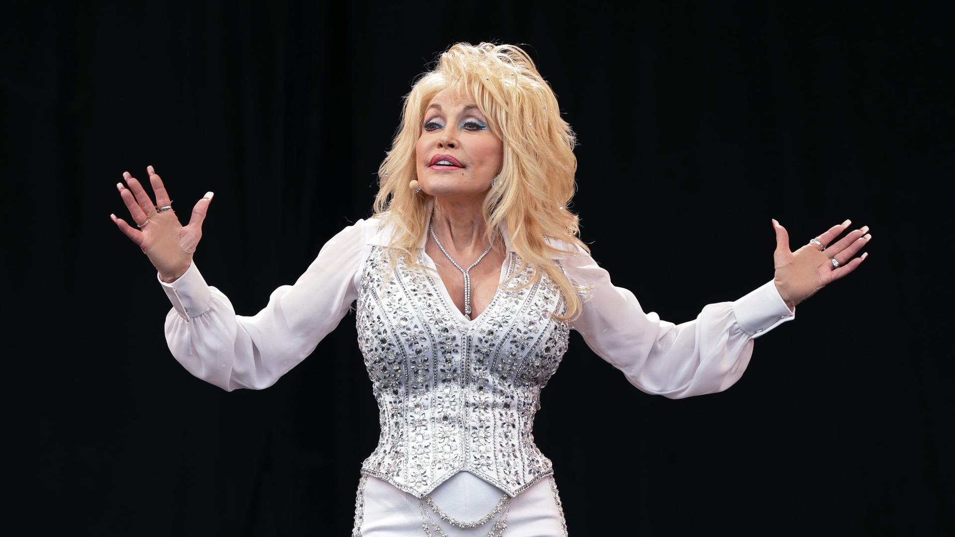 Dolly Parton steht auf einer Bühne und breitet die Arme aus. 