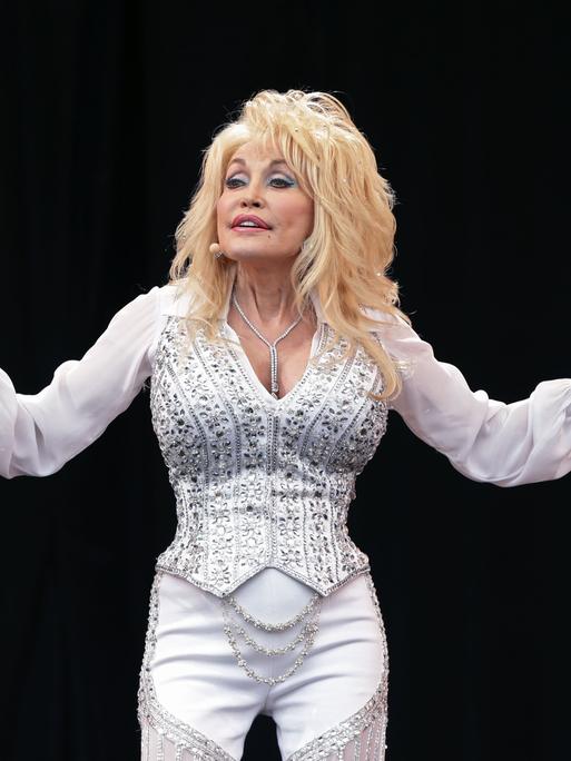 Dolly Parton steht auf einer Bühne und breitet die Arme aus. 