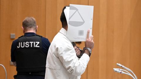 Sulaiman A., Angeklagter, steht mit Handschellen gefesselt im Gerichtssaal und hält sich eine Mappe vor das Gesicht