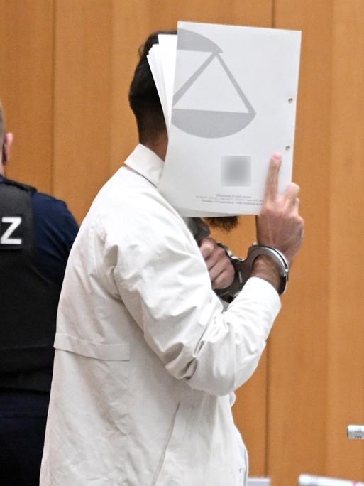 Sulaiman A., Angeklagter, steht mit Handschellen gefesselt im Gerichtssaal und hält sich eine Mappe vor das Gesicht