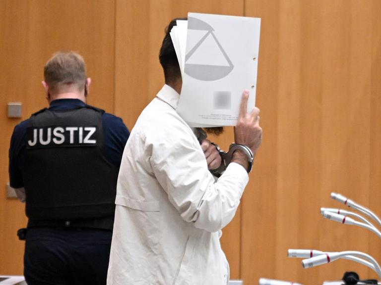 Sulaiman A., Angeklagter, steht mit Handschellen gefesselt im Gerichtssaal und hält sich eine Mappe vor das Gesicht