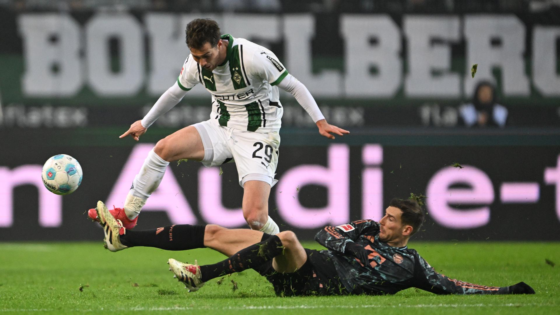 Zu sehen ist eine Spiels-Szene auf einem Fußballfeld. Joe Scally und Leon Goretzka kämpfen um den Ball. Letzterer versucht Ersterem am Boden liegend mit einer Grätsche das Leder abzujagen.