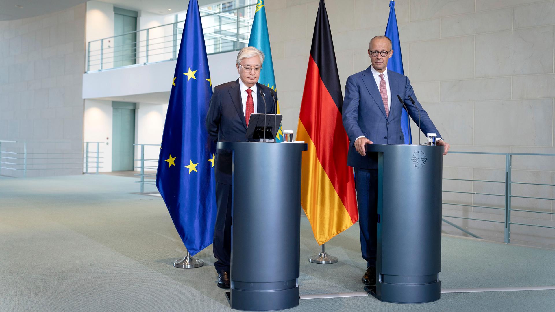 Präsident Tokajew und Bundeskanzler Merz stehen nebeneinander an zwei Rednerpulten, hinter ihnen hängen Fahnen.