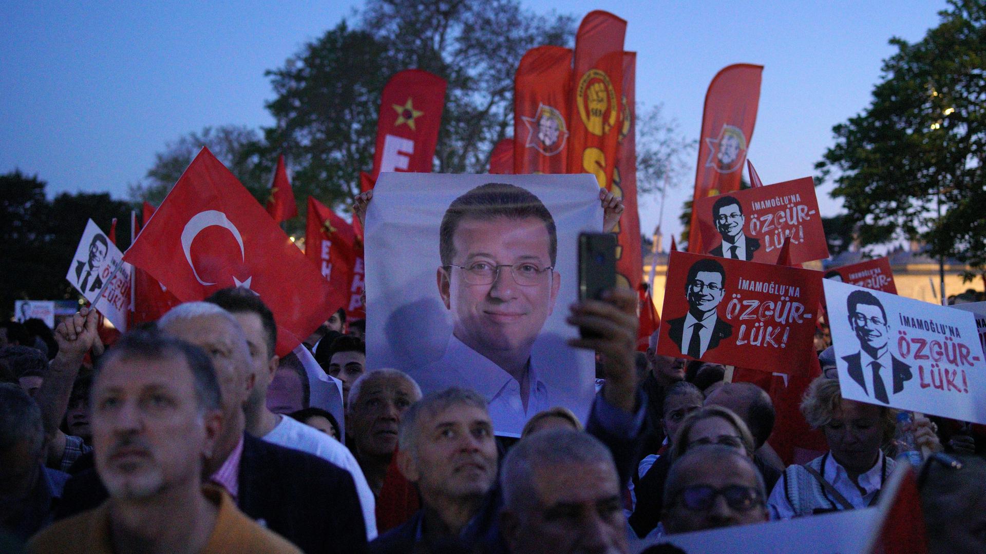 Zahlreiche Menschen stehen zusammen in der Dämmerung. Sie halten unter anderem Türkei-Flaggen und Bilder von Imamoglu hoch. Zahlreiche Menschen stehen zusammen in der Dämmerung. Sie halten unter anderem Türkei-Flaggen und Bilder von Imamoglu hoch.