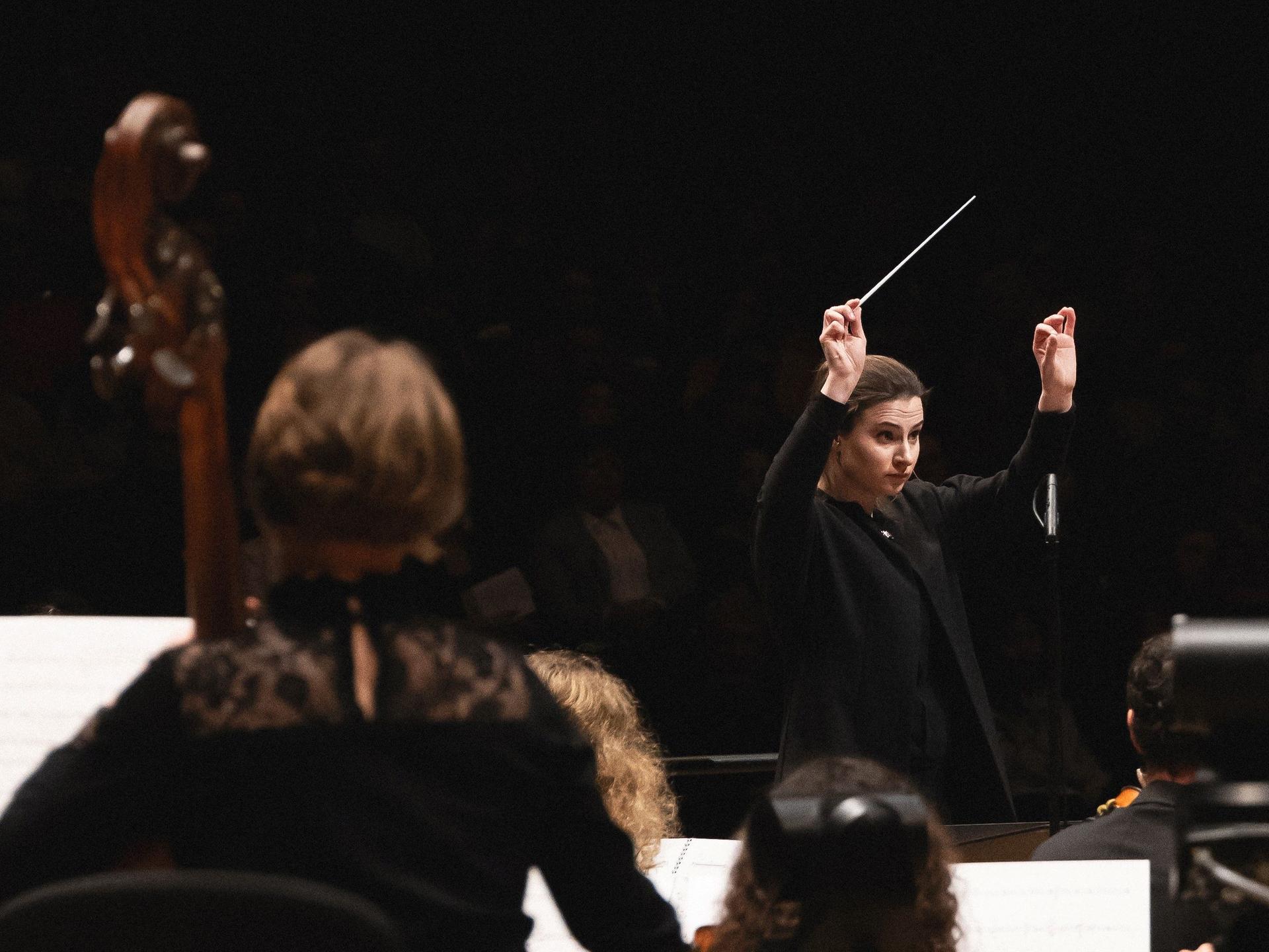 Eine junge Frau mit Pferdeschwanz hebt konzentriert die Arme. In einer Hand hebt sich ihr Dirigierstab von der dunklen Umgebung ab.