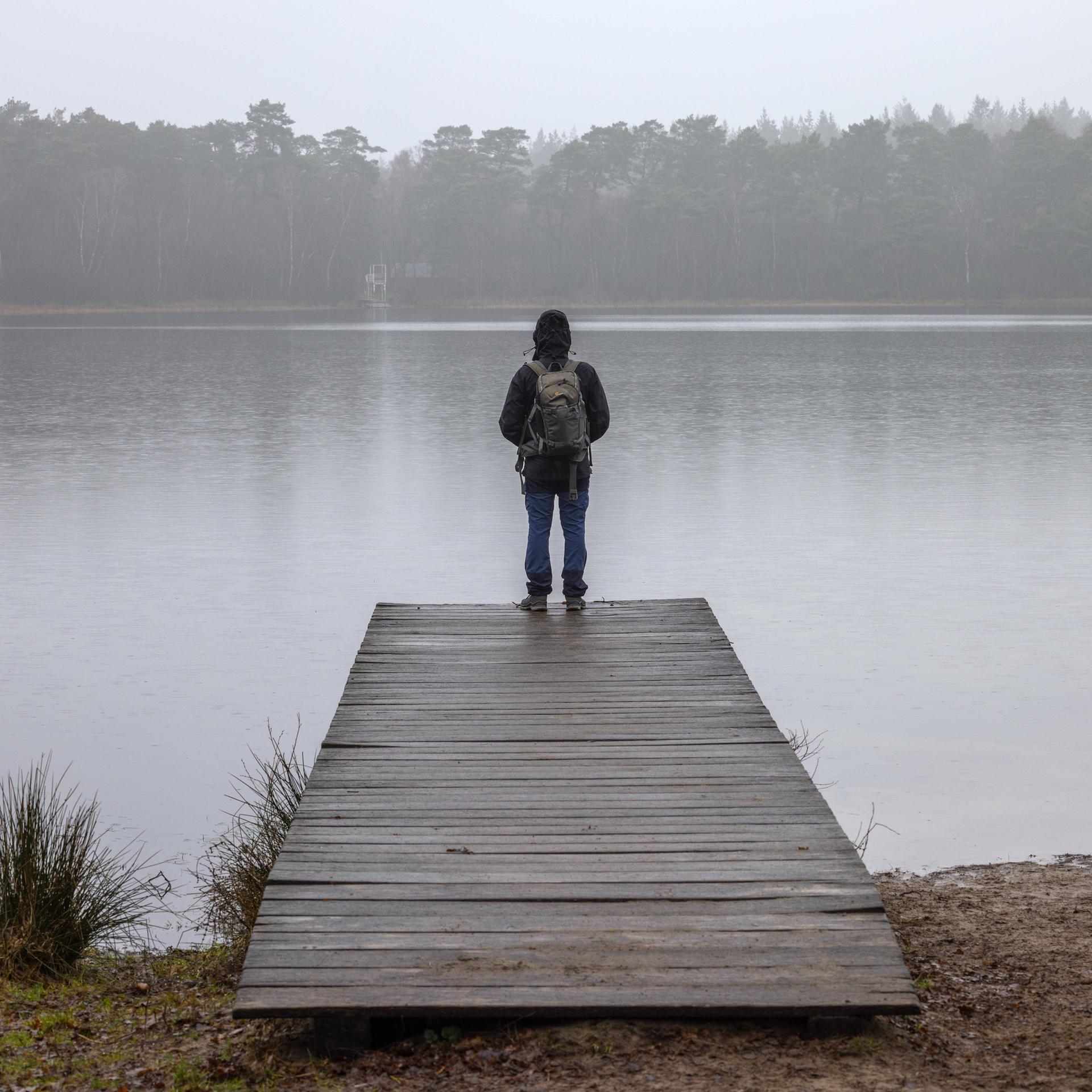 Ein Mann mit Rucksack steht auf einem Holzsteg an einem See vor Wald in einer herbstlich-nebligen Landschaft