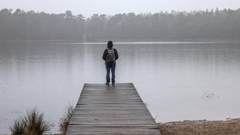 Ein Mann mit Rucksack steht auf einem Holzsteg an einem See vor Wald in einer herbstlich-nebligen Landschaft.