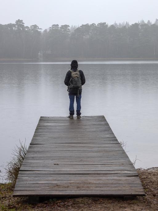 Ein Mann mit Rucksack steht auf einem Holzsteg an einem See vor Wald in einer herbstlich-nebligen Landschaft