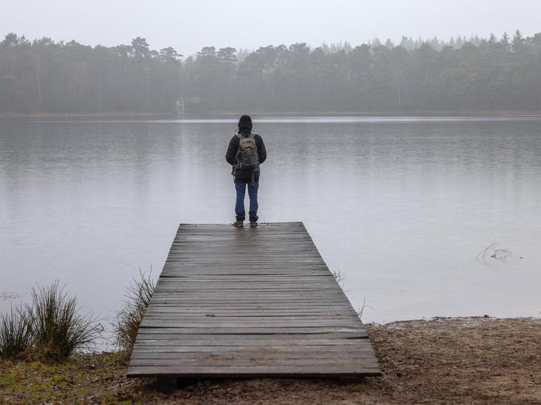 Ein Mann mit Rucksack steht auf einem Holzsteg an einem See vor Wald in einer herbstlich-nebligen Landschaft