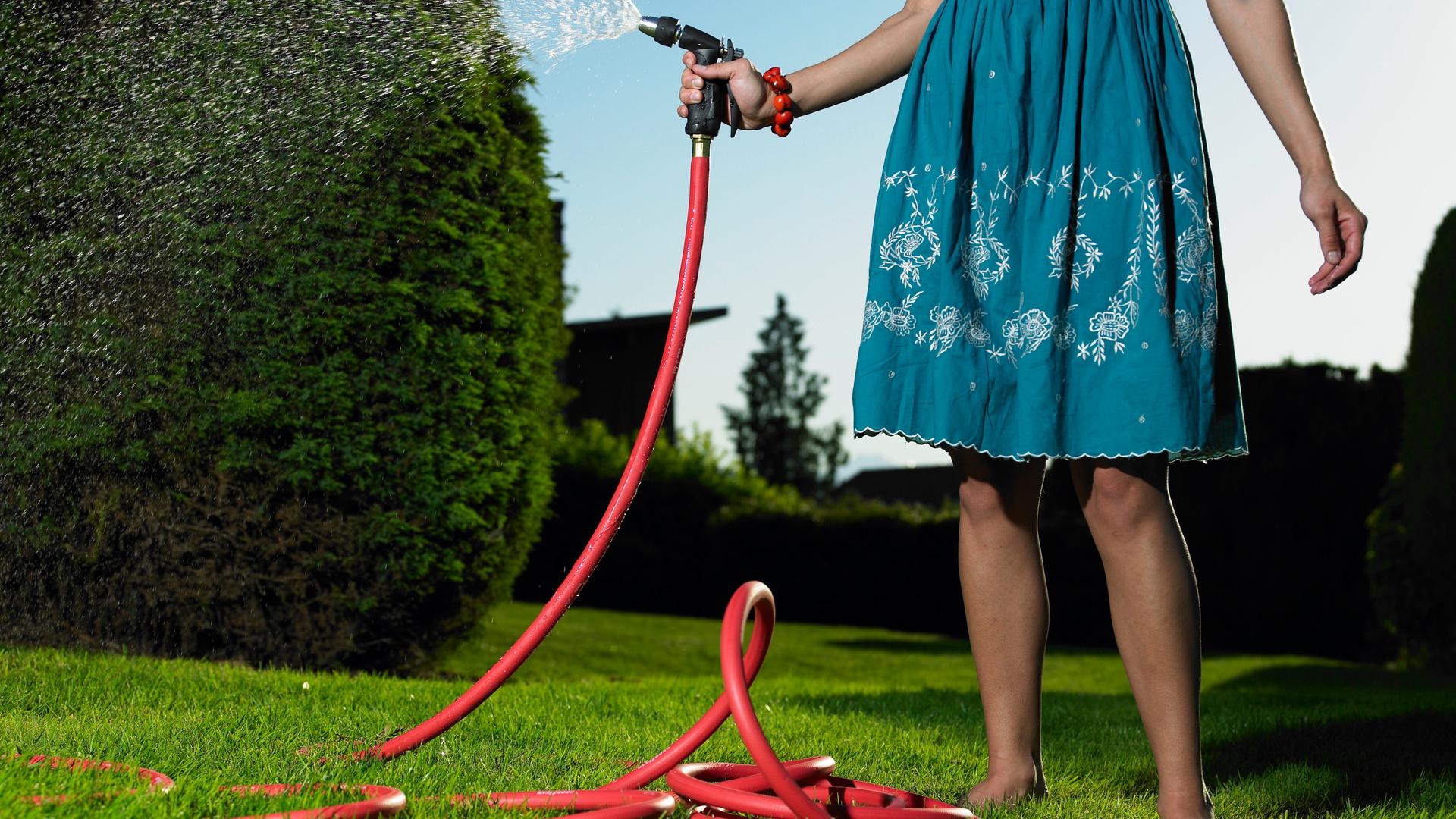 Eine Frau im Sommerkleid bewässert einen Garten mit einem roten Gartenschlauch.