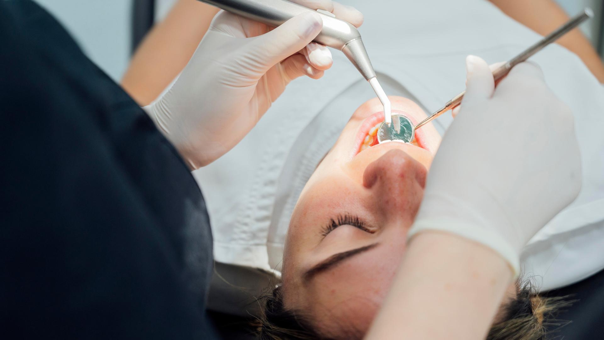 Eine Person liegt auf einem Zahnarztstuhl und wird mit einem Spiegel im Mund untersucht.