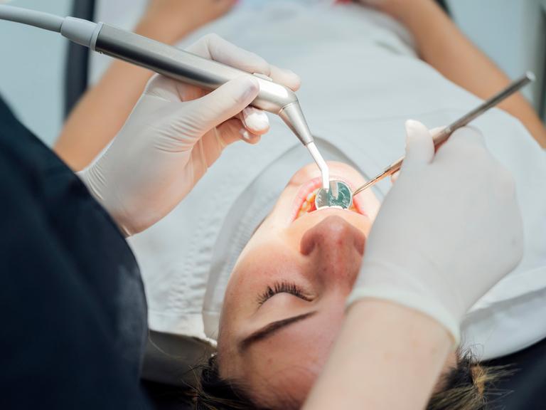 Eine Person liegt auf einem Zahnarztstuhl und wird mit einem Spiegel im Mund untersucht.