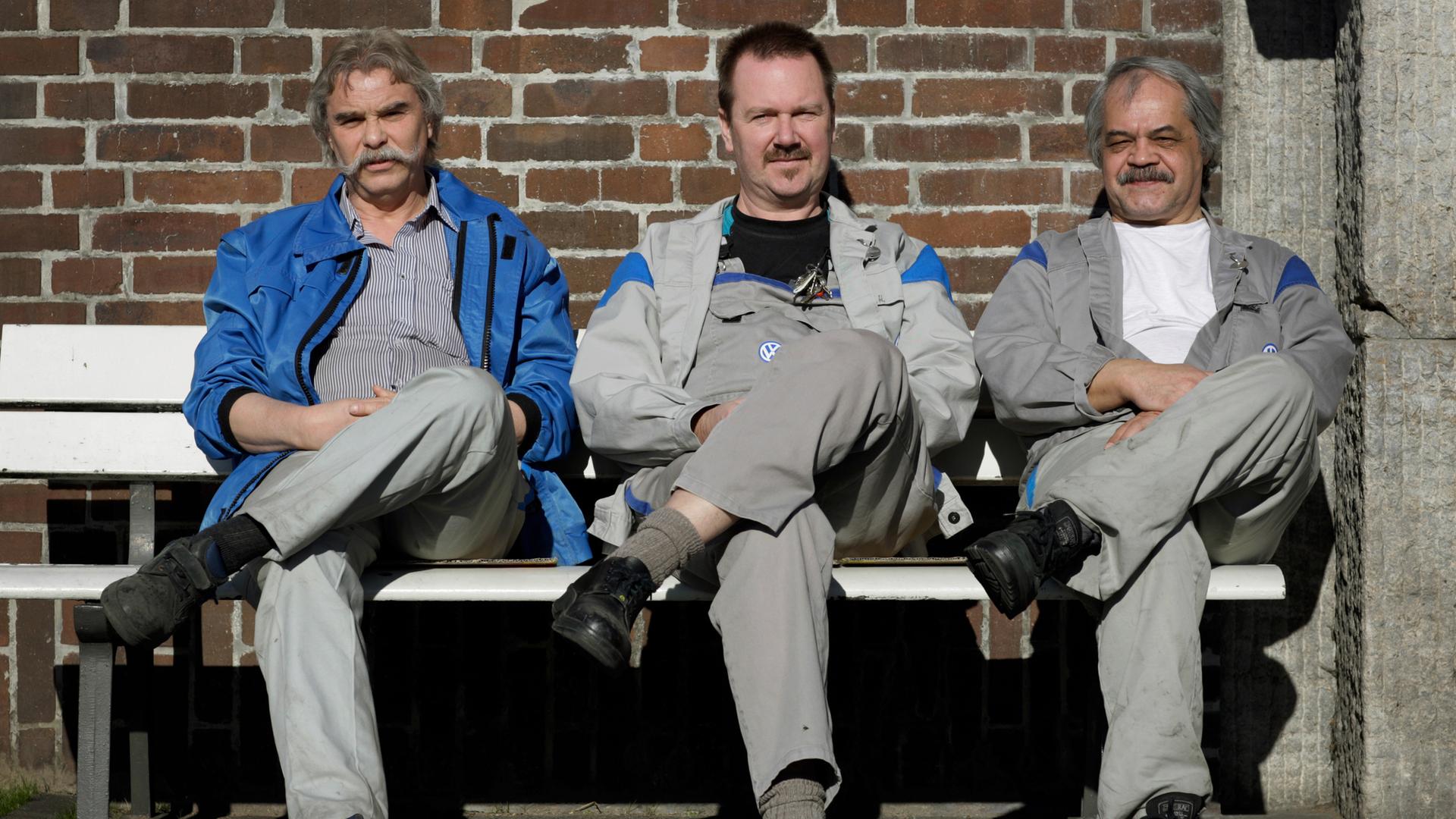VW-Arbeiter bei der Pause: Drei Männer sitzen in Arbeitskleidung auf einer Bank, schlagen das rechte Bein über das linke und blicken in die Kamera.