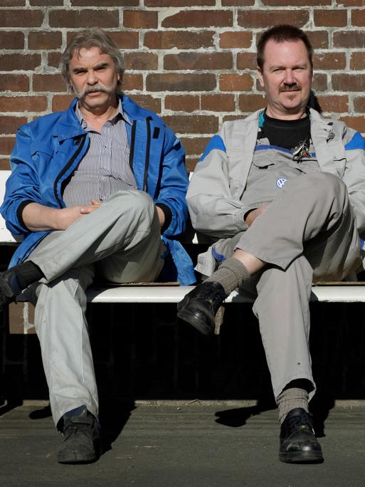 VW-Arbeiter bei der Pause: Drei Männer sitzen in Arbeitskleidung auf einer Bank, schlagen das rechte Bein über das linke und blicken in die Kamera.