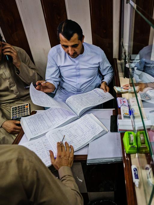 Geldwechsler in Kabul, Afghanistan sitzen in einer Art Wechselstube. Listen werden hin und her gereicht. Es liegen Fernbedienungen und Geldbündel herum.