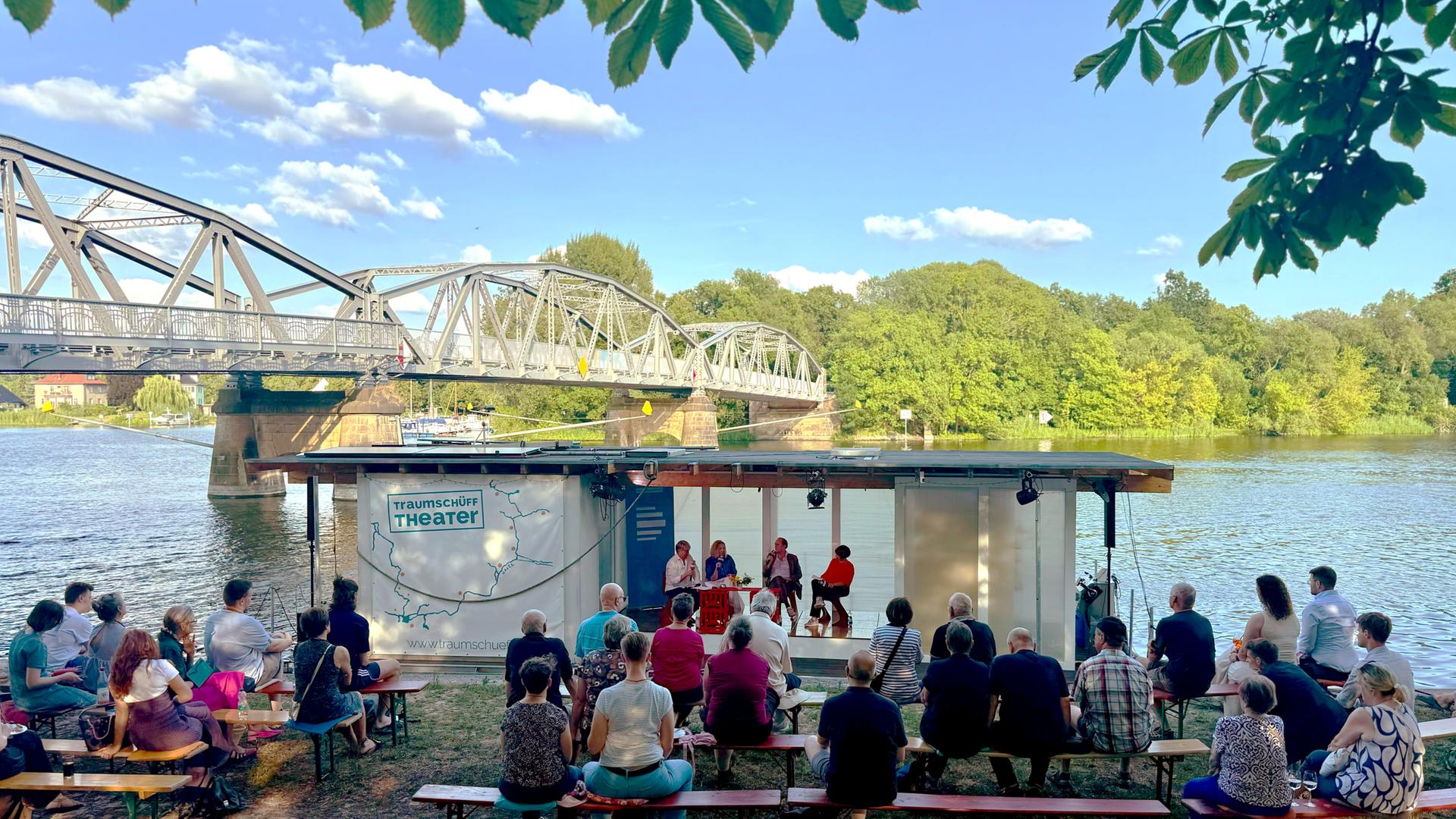 Menschen sitzen auf Bierbänken am Ufer der Havel im Brandenburger Ortsteil Plaue, wo das Theaterschiff Traumschüff anlegt, von dessen Bord der Politikpodcast aufgezeichnet wird. Dahinter eine historische Stahlbrücke. Menschen sitzen auf Bierbänken am Ufer der Havel im Brandenburger Ortsteil Plaue, wo das Theaterschiff Traumschüff anlegt, von dessen Bord der Politikpodcast aufgezeichnet wird. Dahinter eine historische Stahlbrücke.