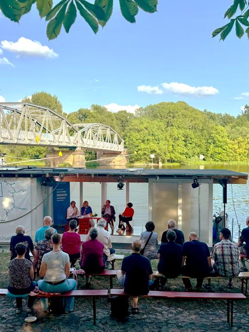 Menschen sitzen auf Bierbänken am Ufer der Havel im Brandenburger Ortsteil Plaue, wo das Theaterschiff Traumschüff anlegt, von dessen Bord der Politikpodcast aufgezeichnet wird. Dahinter eine historische Stahlbrücke.