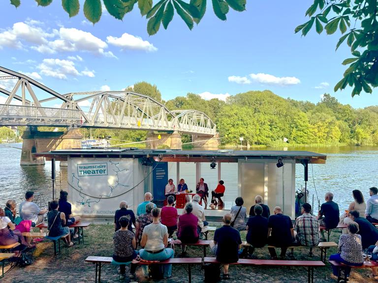 Menschen sitzen auf Bierbänken am Ufer der Havel im Brandenburger Ortsteil Plaue, wo das Theaterschiff Traumschüff anlegt, von dessen Bord der Politikpodcast aufgezeichnet wird. Dahinter eine historische Stahlbrücke.