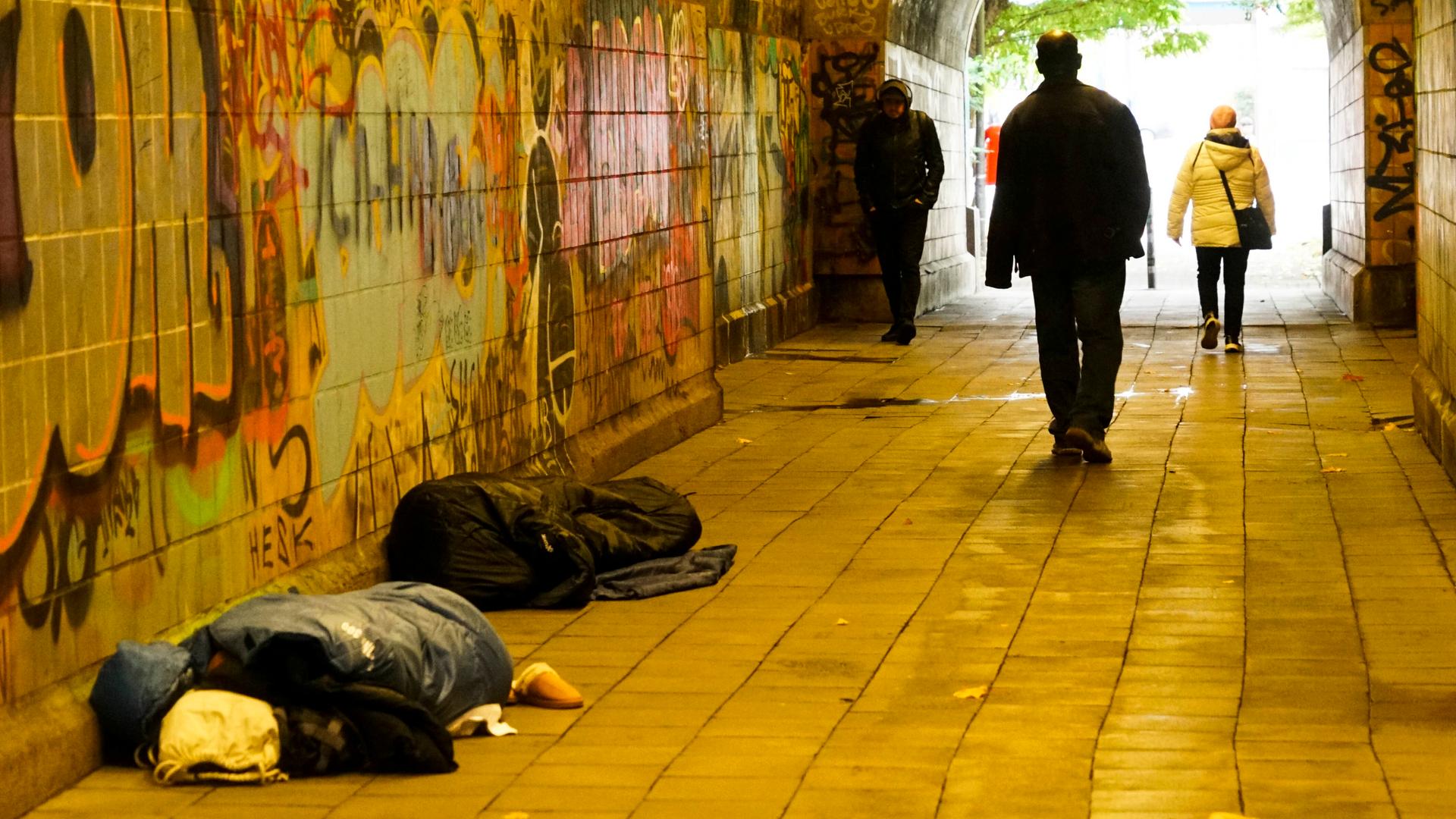 Oblachlose Menschen schlafen am helllichten Tag auf dem Boden mit ihren Habseligkeiten. Passanten gehen an den Menschen auf dem Boden vorbei. Es ist in einem Fußgängertunnel in Hamburg.