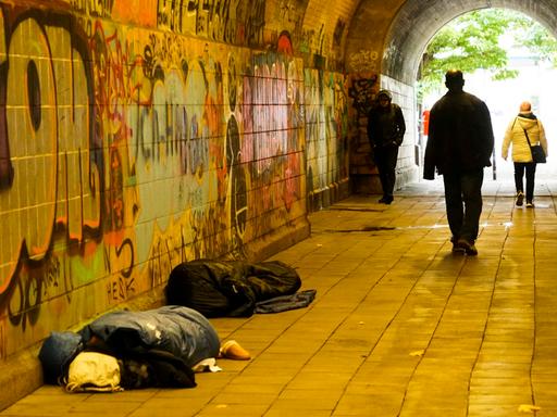 Oblachlose Menschen schlafen am helllichten Tag auf dem Boden mit ihren Habseligkeiten. Passanten gehen an den Menschen auf dem Boden vorbei. Es ist in einem Fußgängertunnel in Hamburg.