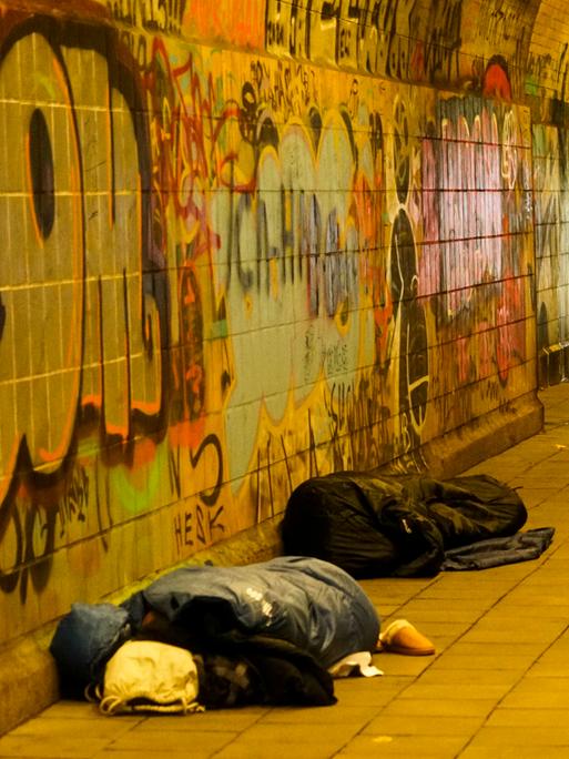 Oblachlose Menschen schlafen am helllichten Tag auf dem Boden mit ihren Habseligkeiten. Passanten gehen an den Menschen auf dem Boden vorbei. Es ist in einem Fußgängertunnel in Hamburg.