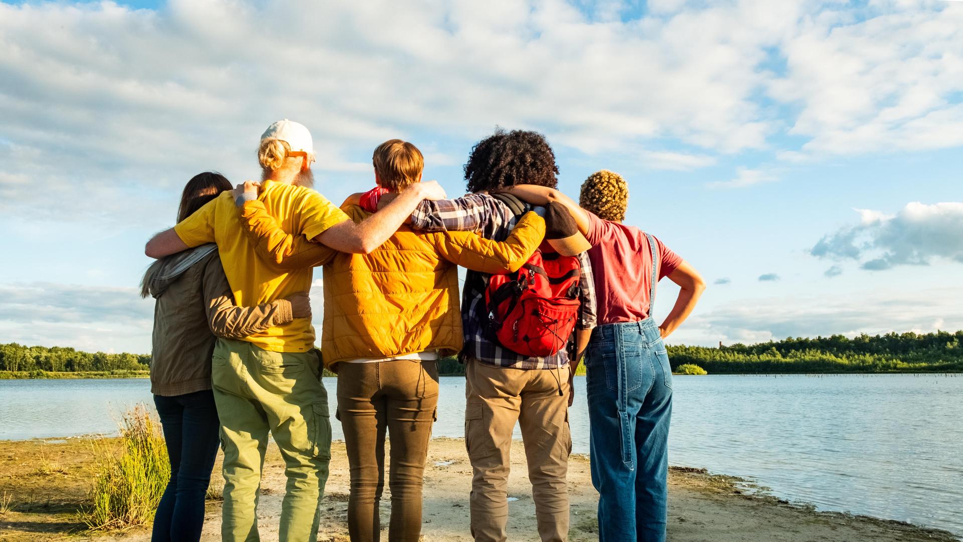 Eine Gruppe junger Freunde stehen mit dem Rücken zum Betrachter vor einem See und halten sich Schulter an Schulter in den Armen. Eine Gruppe junger Freunde stehen mit dem Rücken zum Betrachter vor einem See und halten sich Schulter an Schulter in den Armen.