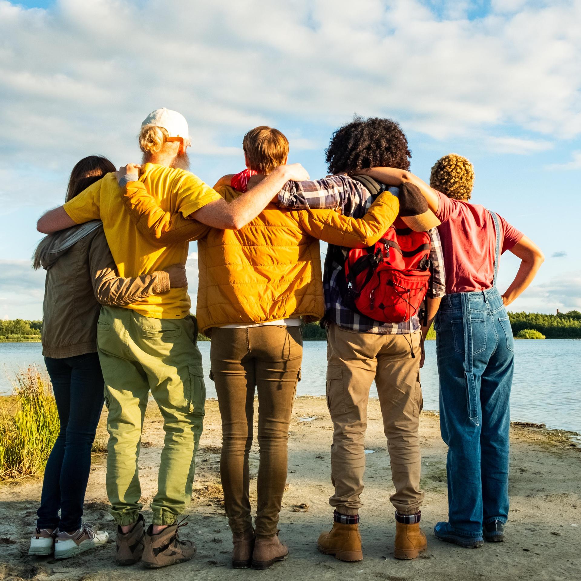 Eine Gruppe junger Freunde stehen mit dem Rücken zum Betrachter vor einem See und halten sich Schulter an Schulter in den Armen.