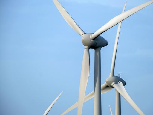 In einem Windpark stehen mehrere Windräder zur Erzeugung von Strom. Dahinter ist hellblauer Himmel zu sehen. 