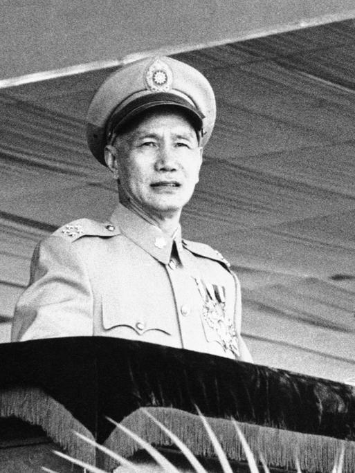 Das Foto zeigt Chiang Kai-shek in Militäruniform und Hut auf einem Schwarzweißfoto. Er steht an einem Rednerpult. Vor ihm sind alte Mikrofone aus den 1950er-Jahren zu sehen. 

