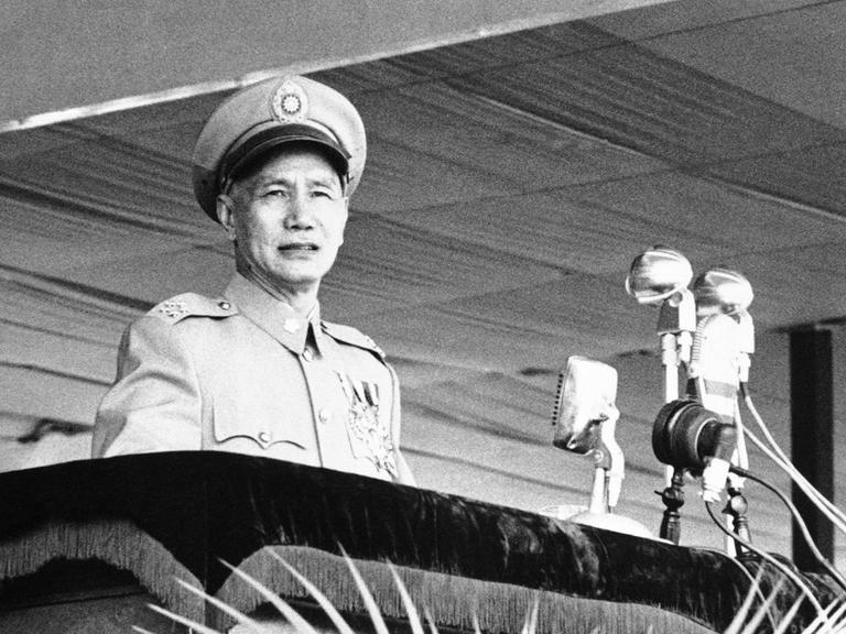 Das Foto zeigt Chiang Kai-shek in Militäruniform und Hut auf einem Schwarzweißfoto. Er steht an einem Rednerpult. Vor ihm sind alte Mikrofone aus den 1950er-Jahren zu sehen. 
