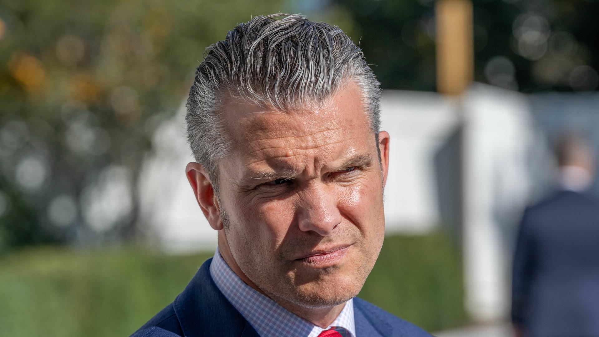 Nahaufnahme des US-Verteidigungsministers Pete Hegseth in blauem Anzug und roter Krawatte. Er kneift seine von der Sonne geblendeten Augen zusammen.
