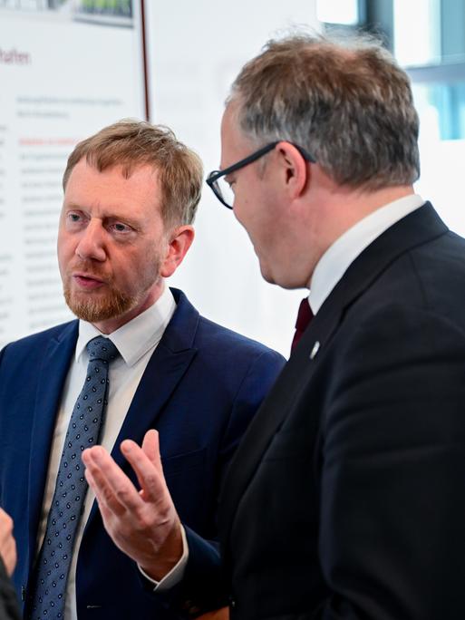 Drei Männer in dunklen Anzügen stehen in einem hellen Konferenzraum eng beieinander und sprechen miteinander. 