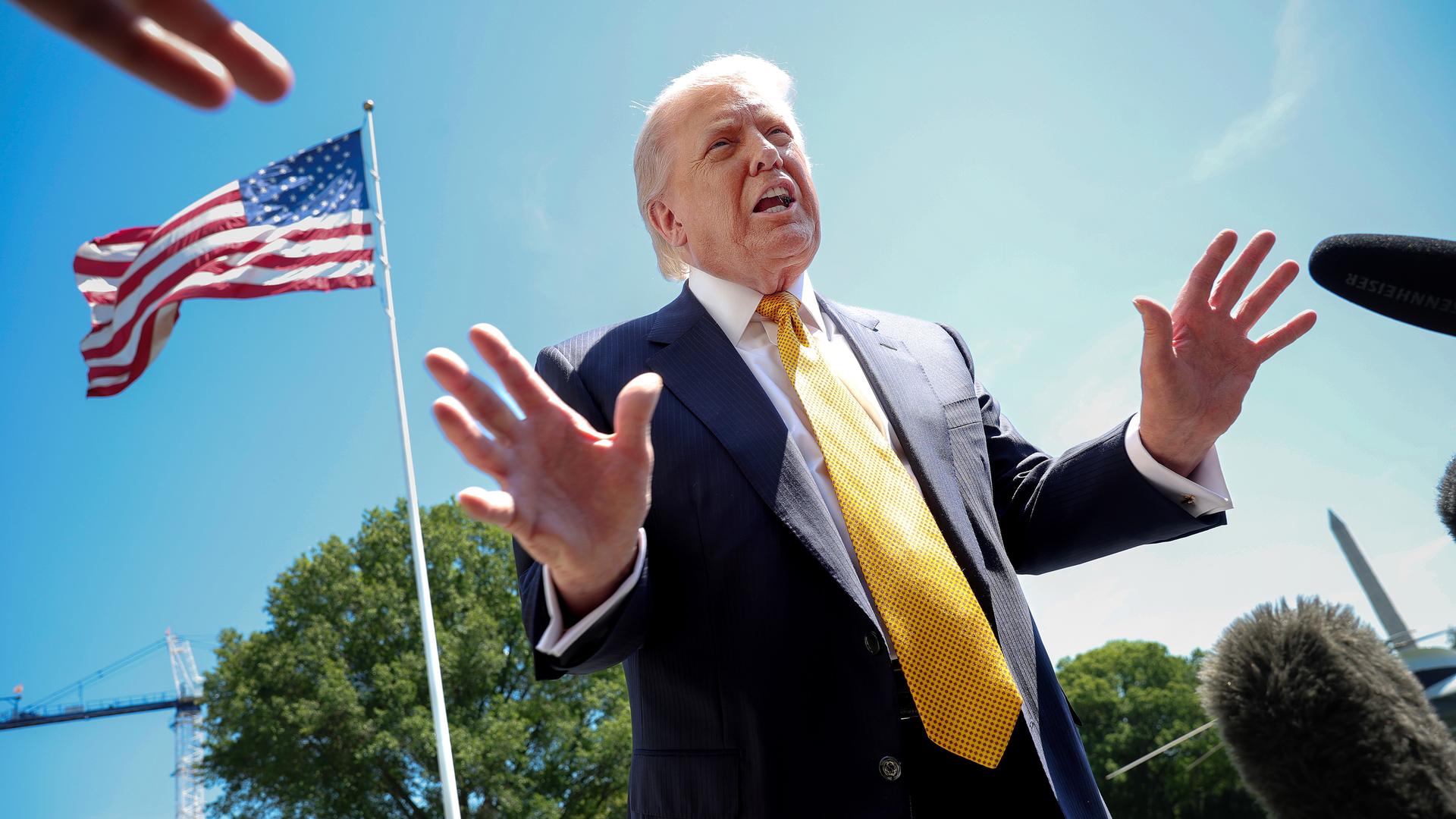US-Präsident Donald Trump spricht mit Journalisten. Das Foto ist von schräg unten aufgenommen. Im Hintergrund ist eine US-Flagge und blauer Himmel zu sehen.