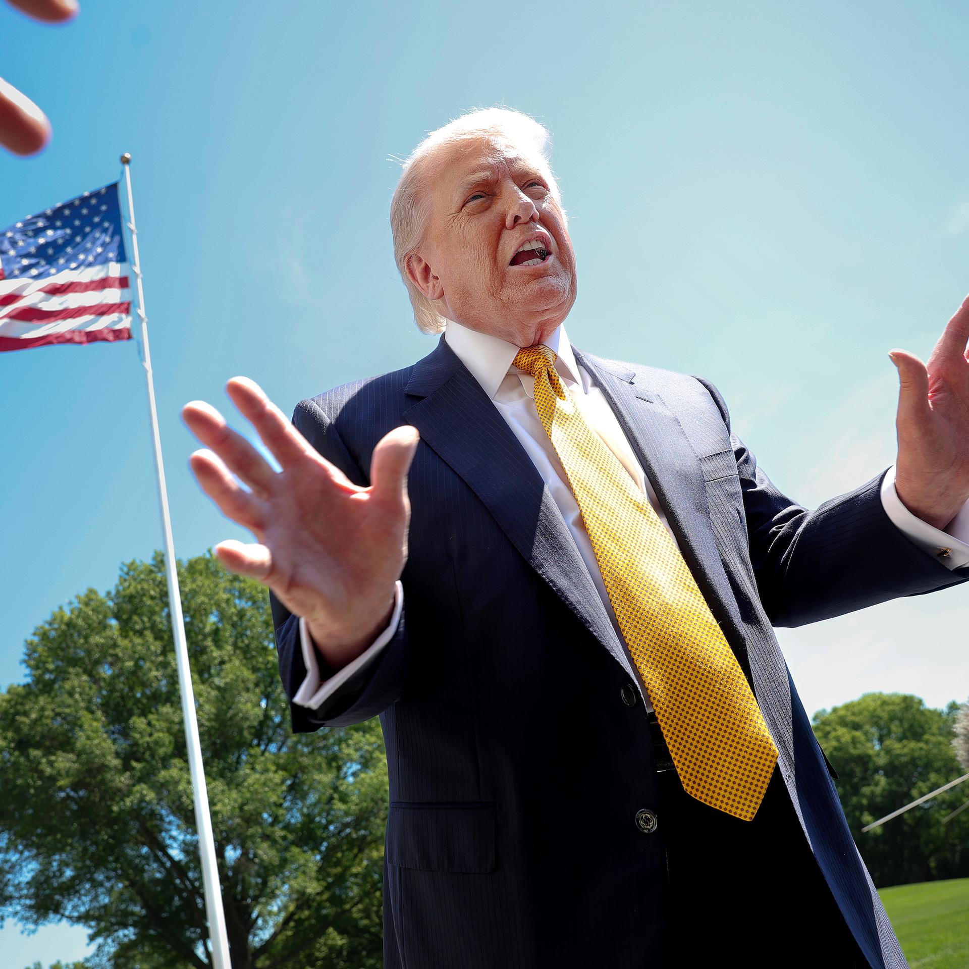 Der US-Präsident Donald Trump spricht mit Journalisten. Das Foto ist von schräg unten aufgenommen. Im Hintergrund ist eine US-Flagge und blauer Himmel zu sehen.