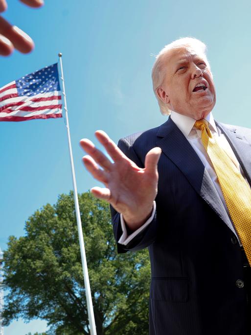 US-Präsident Donald Trump spricht mit Journalisten. Das Foto ist von schräg unten aufgenommen. Im Hintergrund ist eine US-Flagge und blauer Himmel zu sehen. US-Präsident Donald Trump spricht mit Journalisten. Das Foto ist von schräg unten aufgenommen. Im Hintergrund ist eine US-Flagge und blauer Himmel zu sehen.