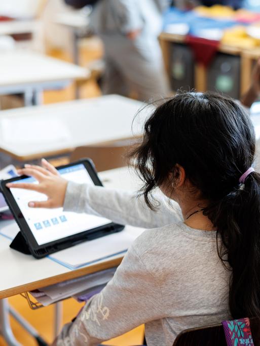 Zwei Mädchen mit dunklen Haaren sitzen an einem Schreibtisch in einem Klassenzimmer und bearbeiten mit Hilfe von ihrem Tablet ein Aufgabenblatt.