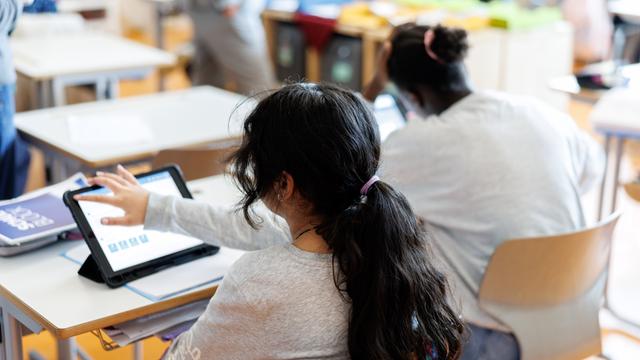 Zwei Mädchen mit dunklen Haaren sitzen an einem Schreibtisch in einem Klassenzimmer und bearbeiten mit Hilfe von ihrem Tablet ein Aufgabenblatt.