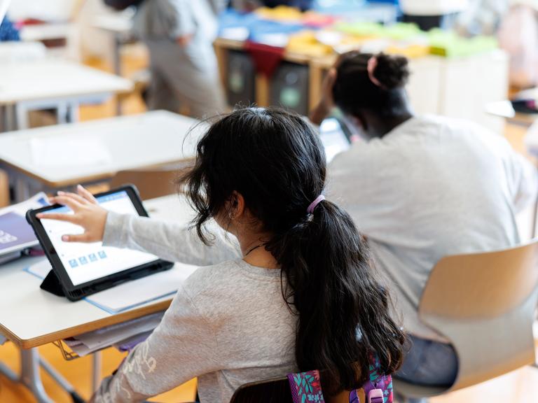 Zwei Mädchen mit dunklen Haaren sitzen an einem Schreibtisch in einem Klassenzimmer und bearbeiten mit Hilfe von ihrem Tablet ein Aufgabenblatt.