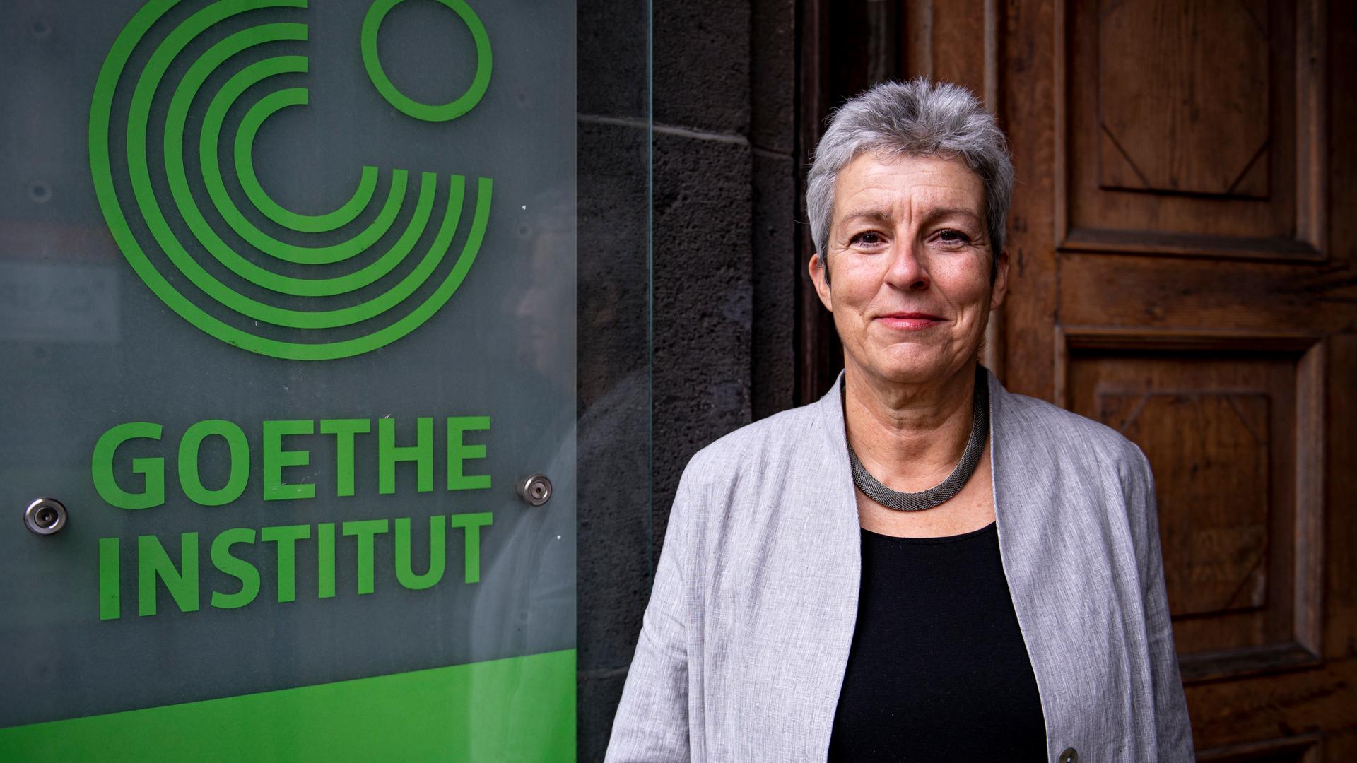 Carola Lentz,  Präsidentin des Goethe-Instituts, mit grauem Kurzhaarschnitt, in hellgrauem Sakko, steht neben dem Türschild des Goethe-Instituts in Berlin.