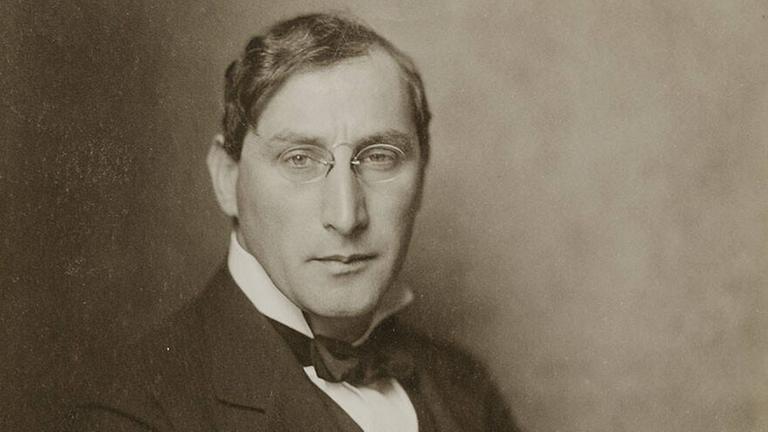 Eine historische Fotographie zeigt einen gepflegten Mann mit Seitescheitel, kleiner Brille und Tuch im hoch aufgestellten Kragen.