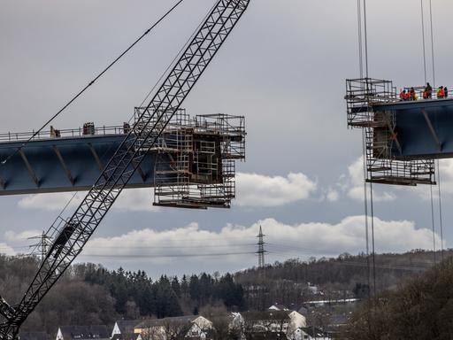 Untersicht auf zwei Brückenelemente, die per Kran zusammengeführt werden. Am unteren Bildrand ist eine Ortschaft zu sehen. Untersicht auf zwei Brückenelemente, die per Kran zusammengeführt werden. Am unteren Bildrand ist eine Ortschaft zu sehen.